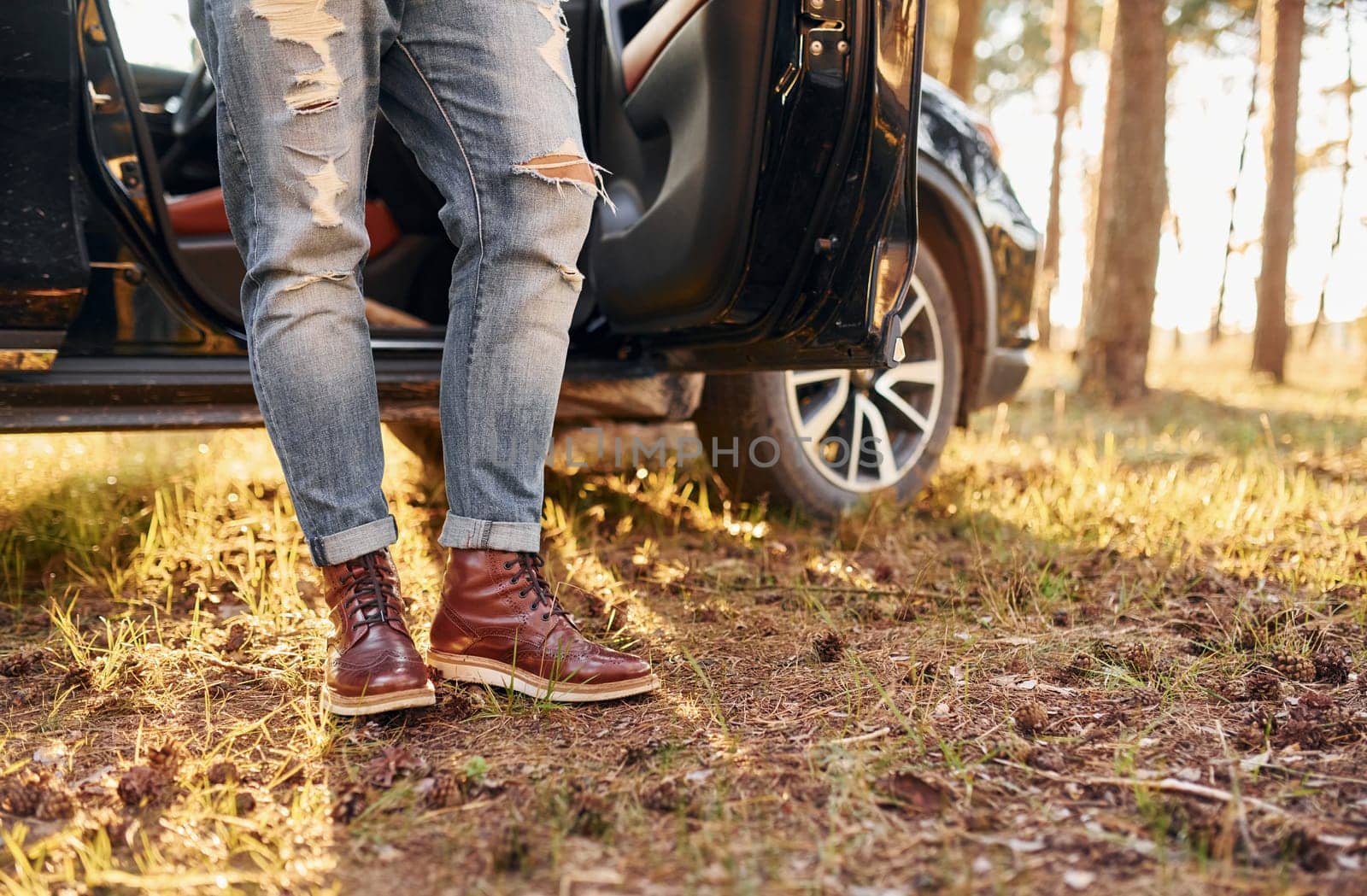 View of jeans and shoes. Man in jeans is outdoors in the forest with his black colored automobile.