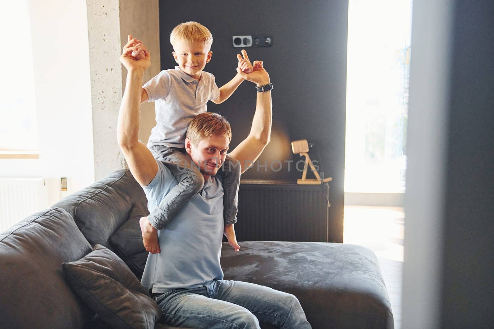 Boy is sitting on the man's shoulders. Father and son is indoors at home together.