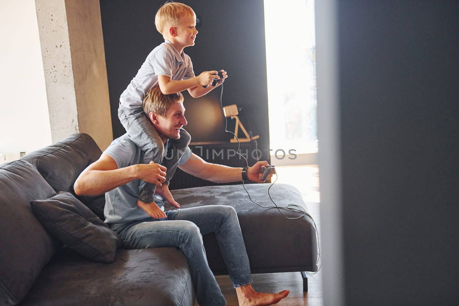 Boy is sitting on the man's shoulders. Father and son is indoors at home together.