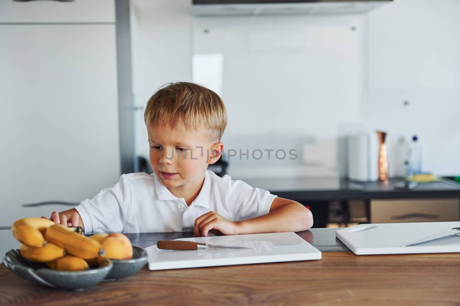 Boy in casual clothes is sitting in the kitchen with bananas on table.