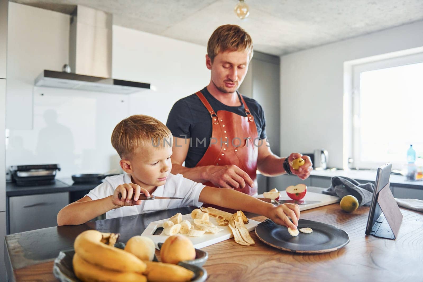 Man is in the apron on kitchen. Father and son is indoors at home together.