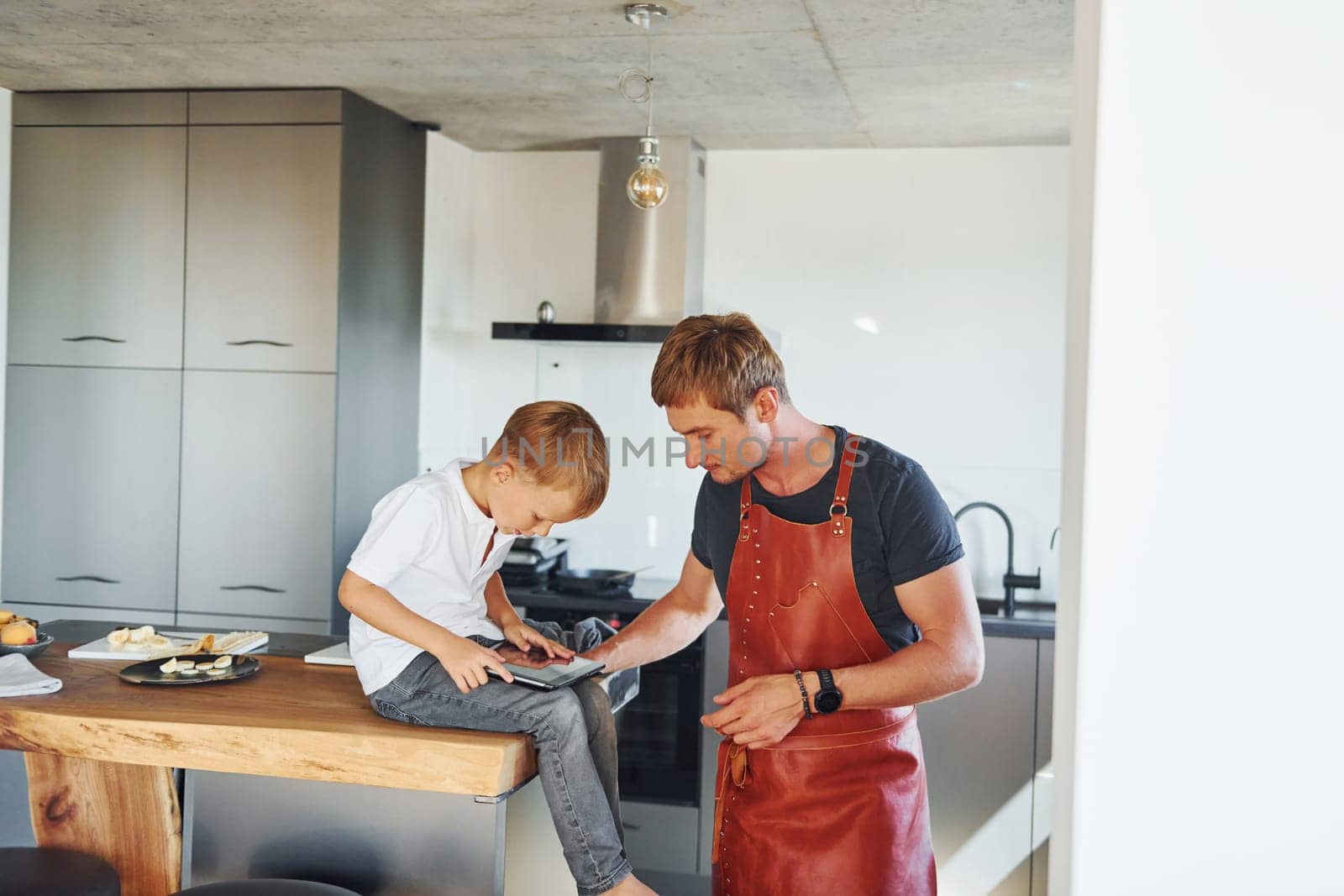 Man is in the apron on kitchen. Father and son is indoors at home together.