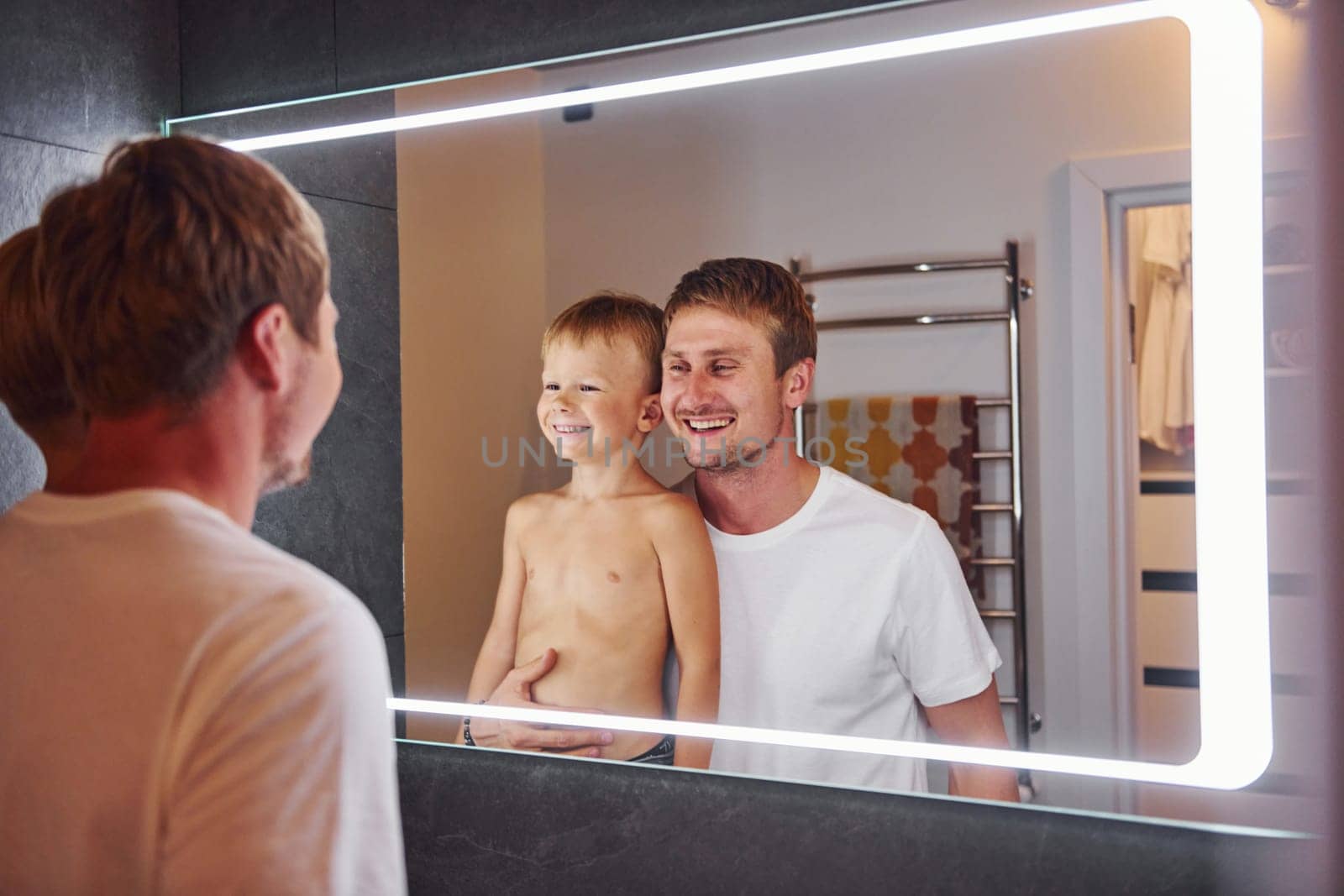 Looking in the mirror in bathroom. Father and son is indoors at home together by Standret