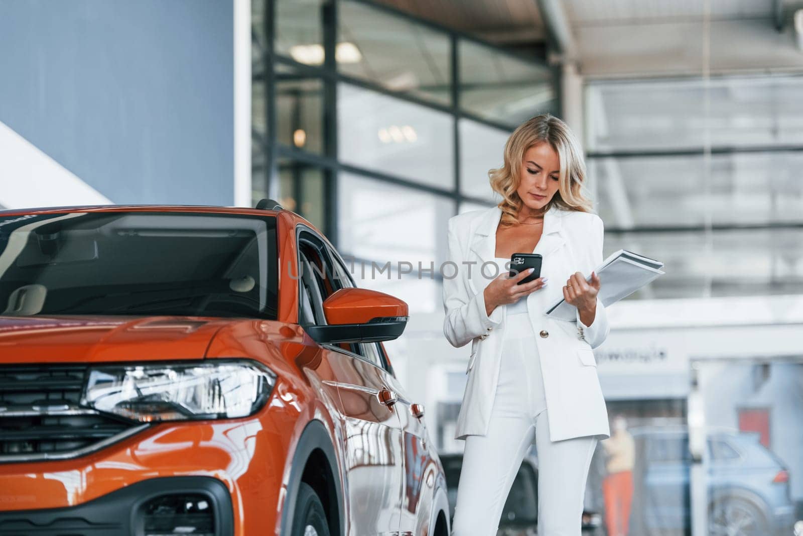 Holding phone. Woman in formal clothes is indoors in the autosalon.