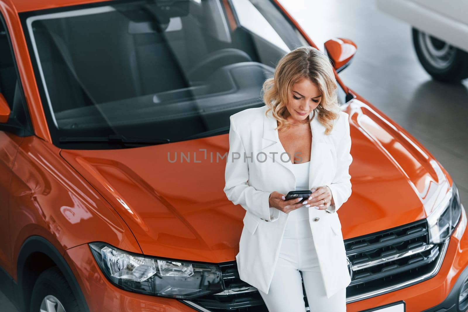 With phone in hand. Woman in formal clothes is indoors in the autosalon.