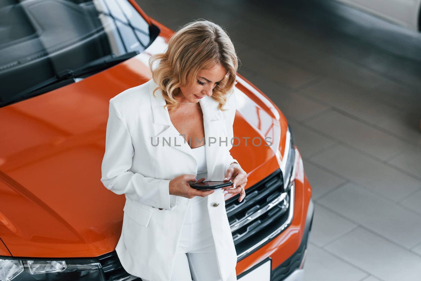 With phone in hand. Woman in formal clothes is indoors in the autosalon.
