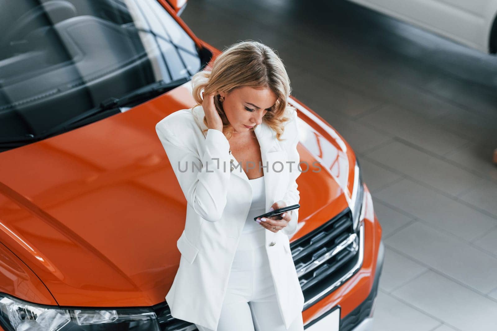Leaning on the car. Woman in formal clothes is indoors in the autosalon.