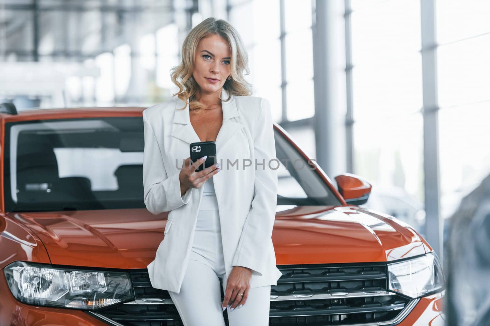 Leaning on the car. Woman in formal clothes is indoors in the autosalon.