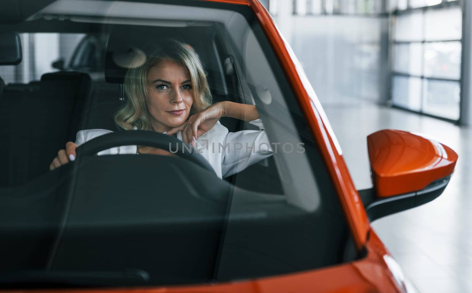Sitting in the car. Woman in formal clothes is indoors in the autosalon.