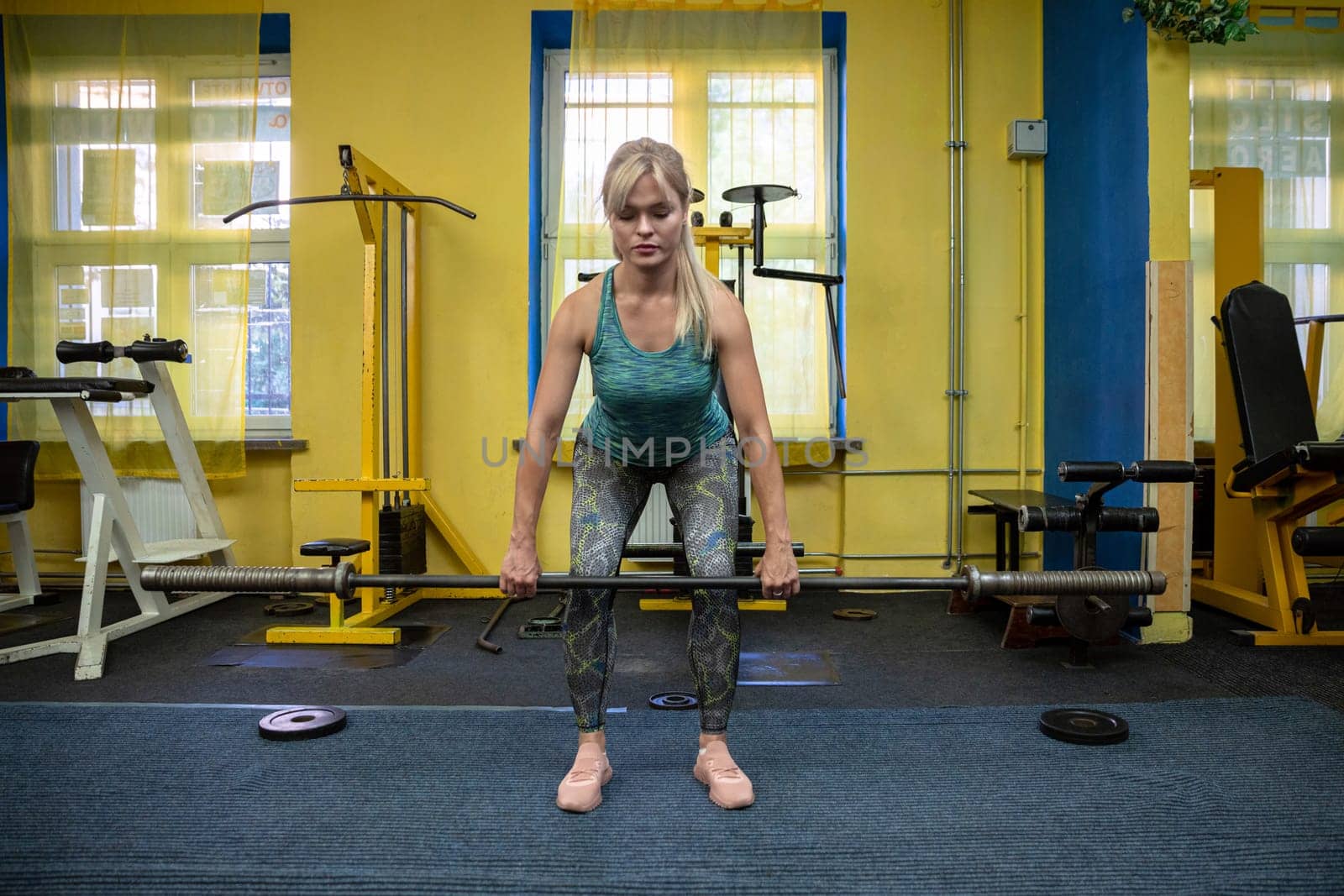 A young woman exercises at the gym by lifting a barbell to knee height. Using sports equipment at the gym. Behind the woman you can see the walls painted yellow and the windows