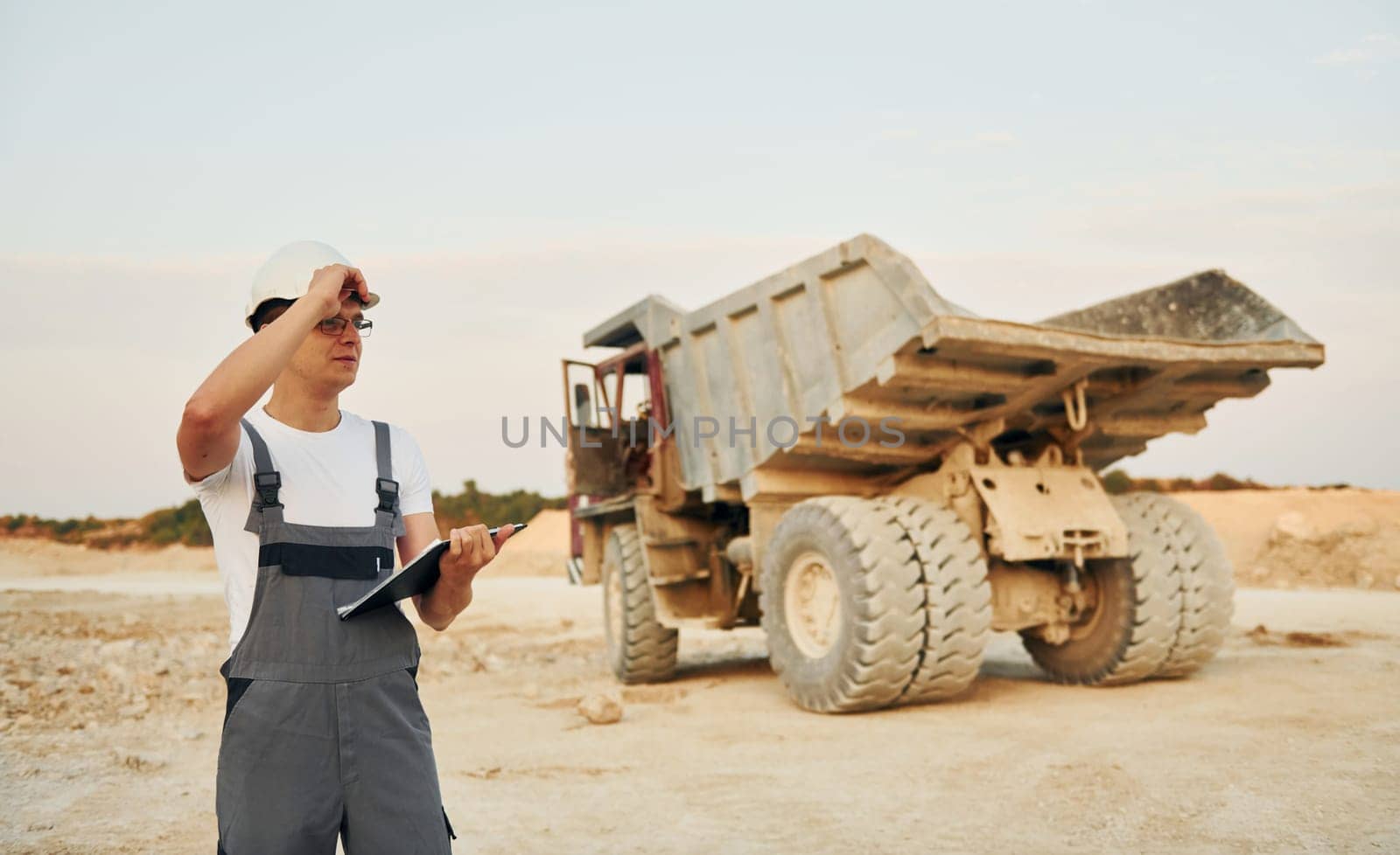 In white hard hat. Worker in professional uniform is on the borrow pit at daytime.