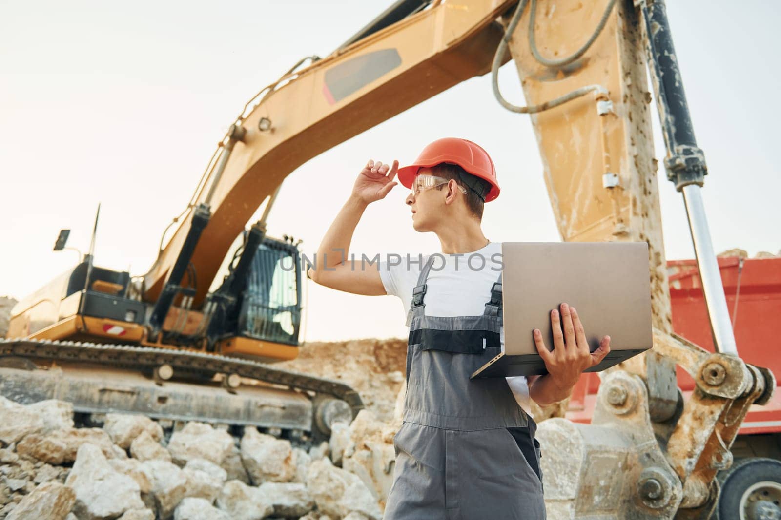 Standing against yellow vehicle. Worker in professional uniform is on the borrow pit at daytime.