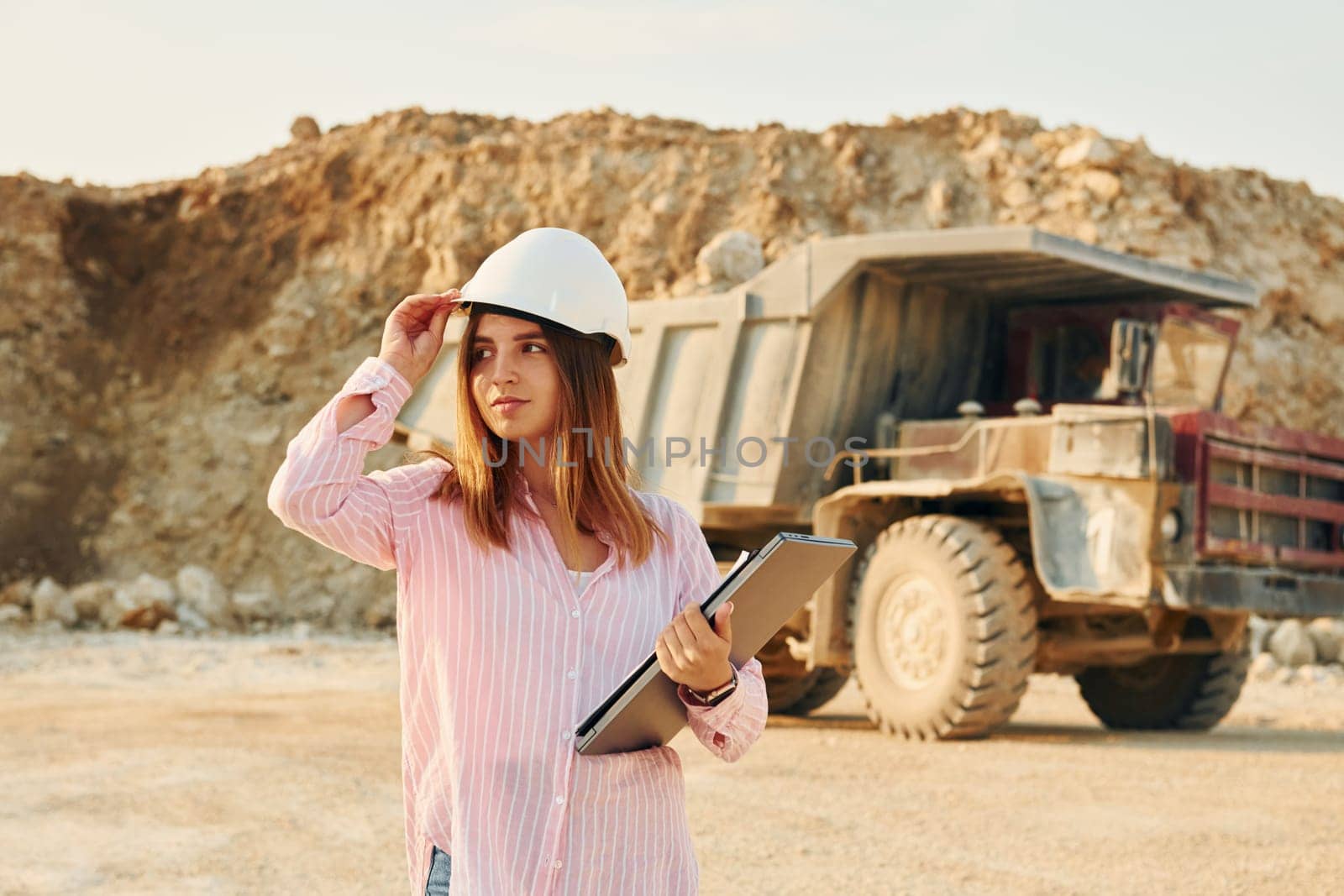 With documents. Young woman with documents in hard hat is standing on the borrow pit.