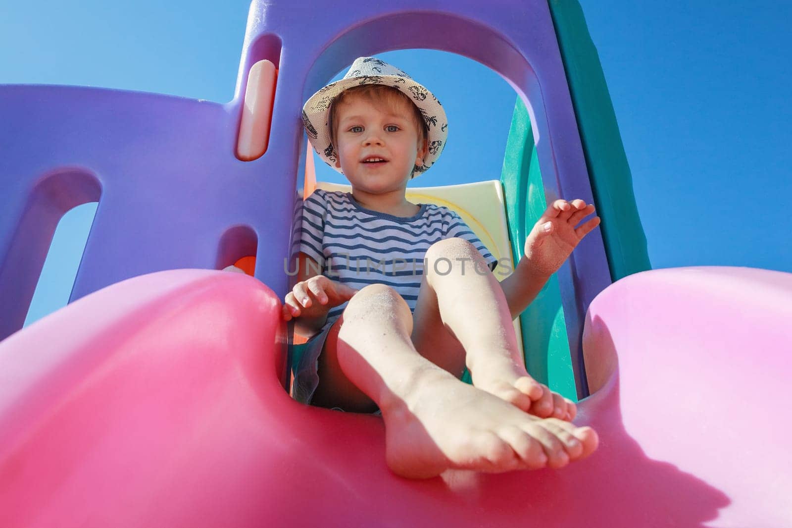 A portrait of a little boy in a striped shirt on a pink slider against a blue sky background. by Lincikas