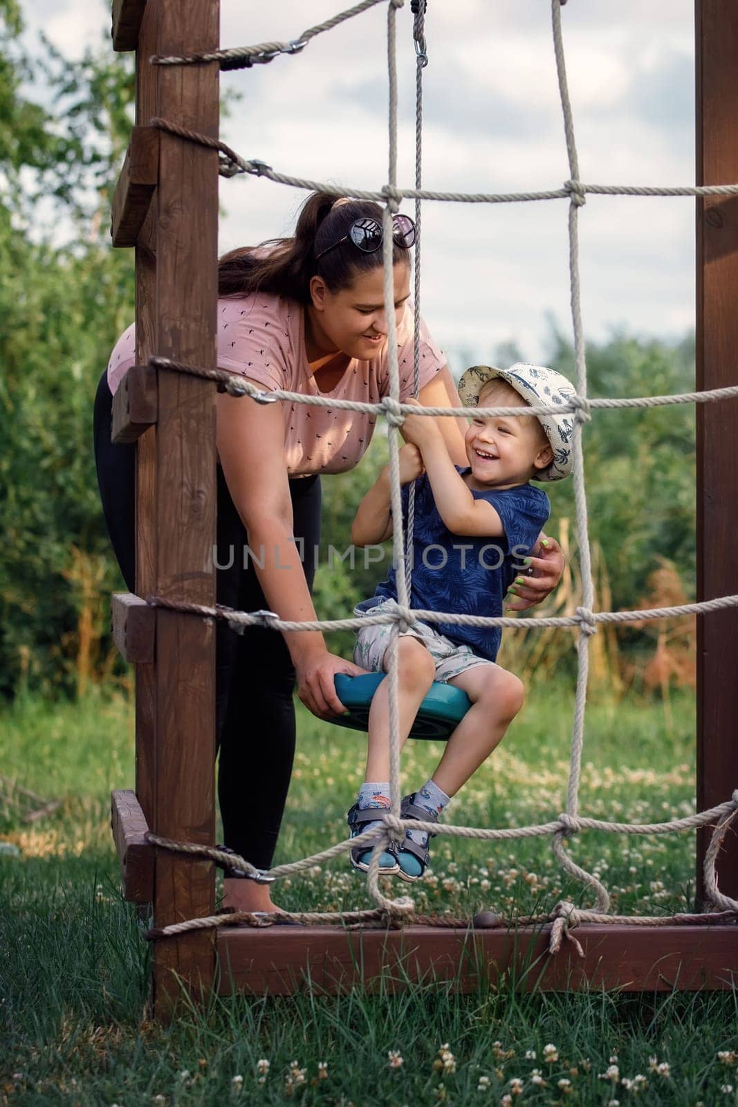 Mom has fun playing with her son on the ropes ladder in the garden playground.