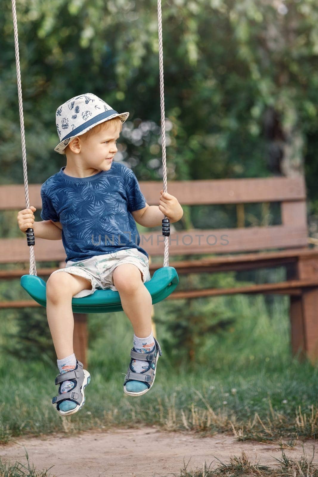 Little child blond boy having fun on a swing outdoor. Summer playground. Child swinging carefully and looks to distance.