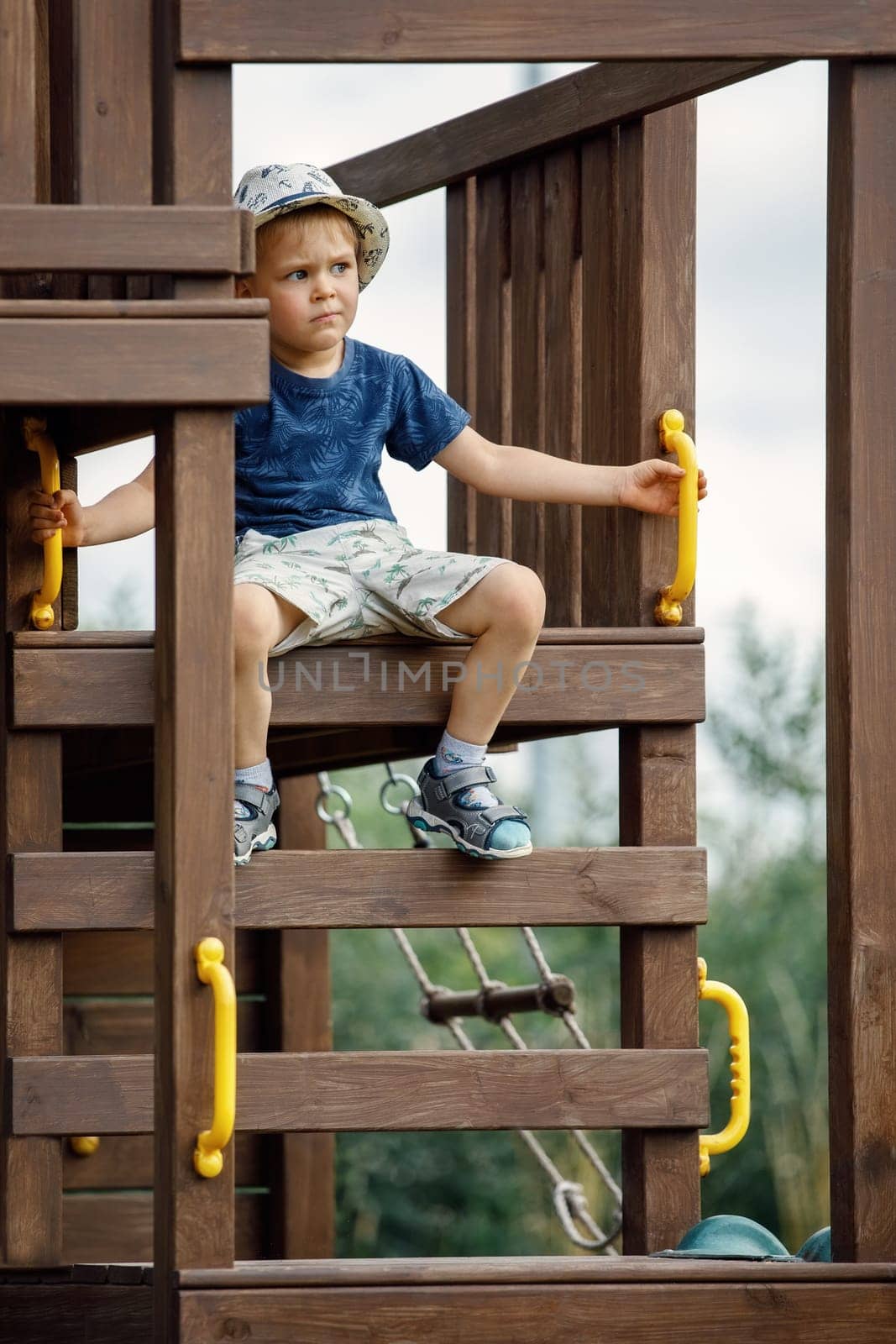 A cute little brave boy sitting at the top of the wooden playground. by Lincikas