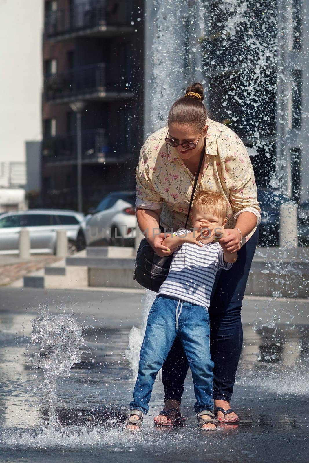 Mom with her son in the city by the water fountain. The little child is frightened by the sudden jets of water. Kids fun in the city on a hot summer day.