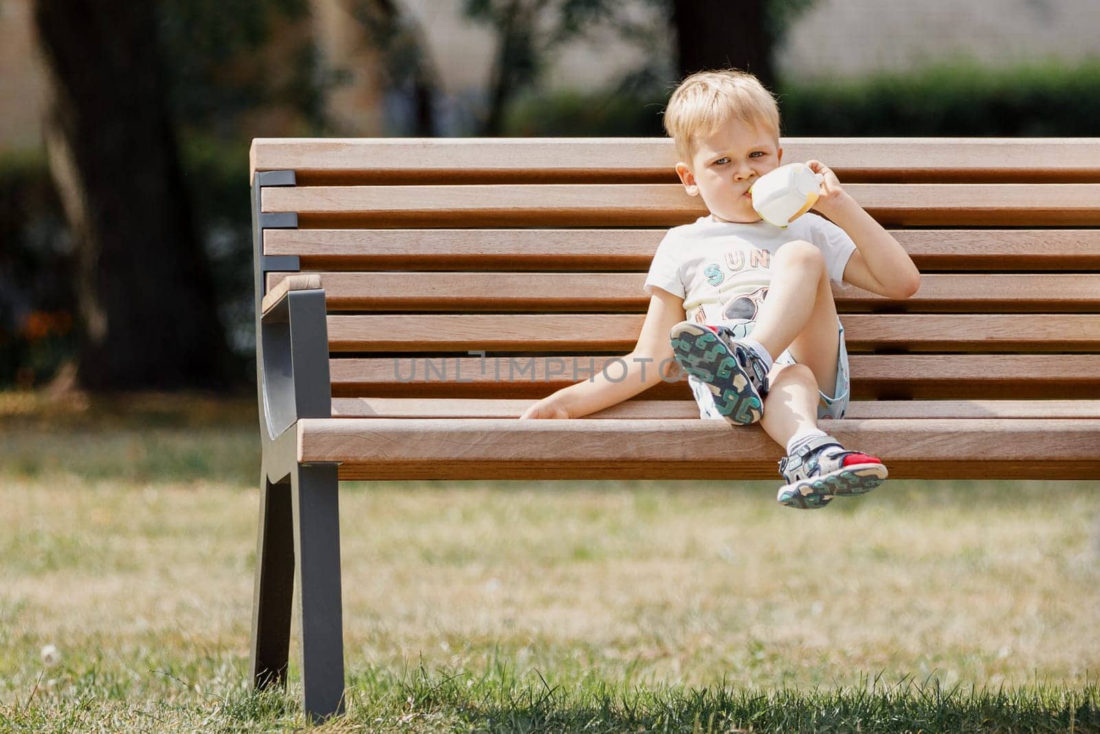 Cute little boy sitting on a bench and drinks juice. by Lincikas