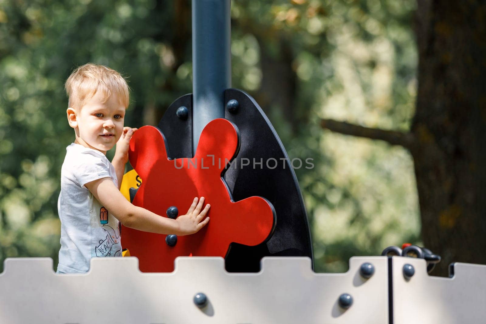 A child is driving the red helm of a ship on a playground. Dark green park tree foliage background. by Lincikas