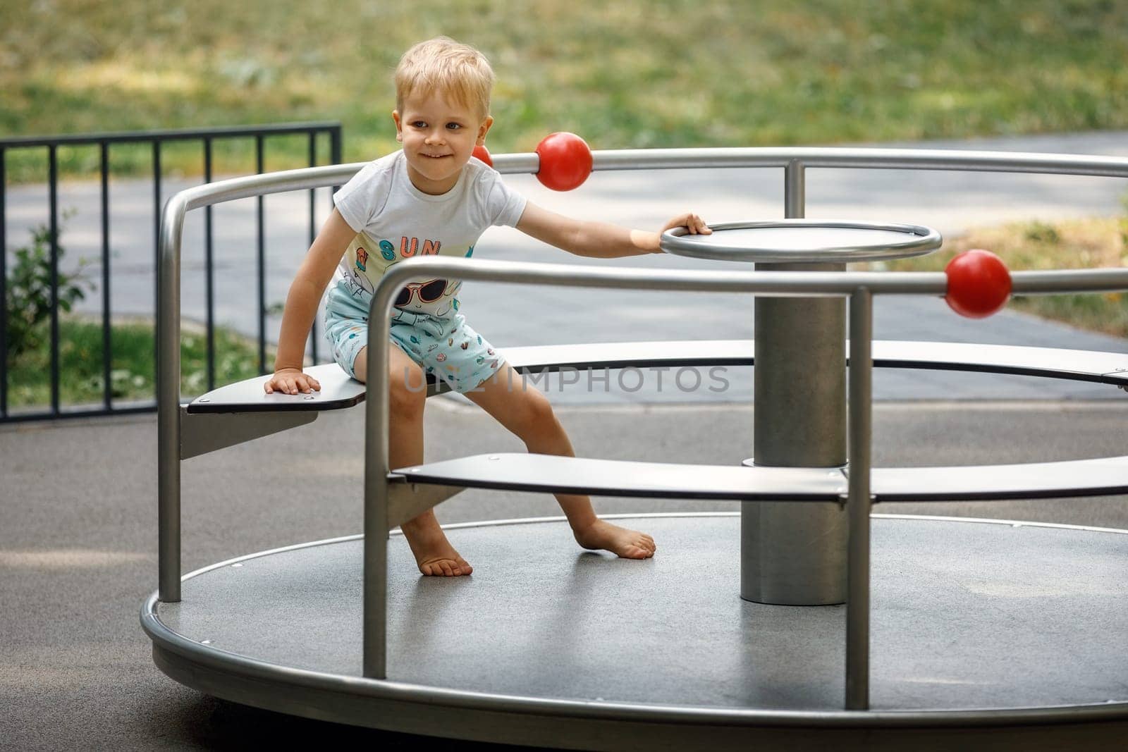 A little boy sits in a metal carousel for children in summer in the park and poses for the camera. by Lincikas