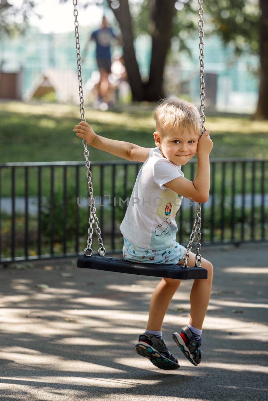 A mischievous little boy swings in a park on a chain swing, the child turns to the camera and smiles happily by Lincikas