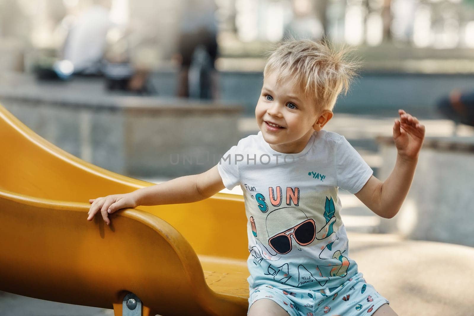 A cheerful, smiling, likeable little boy on the playground on a yellow slider. The child's hair is electrified and bristling by Lincikas