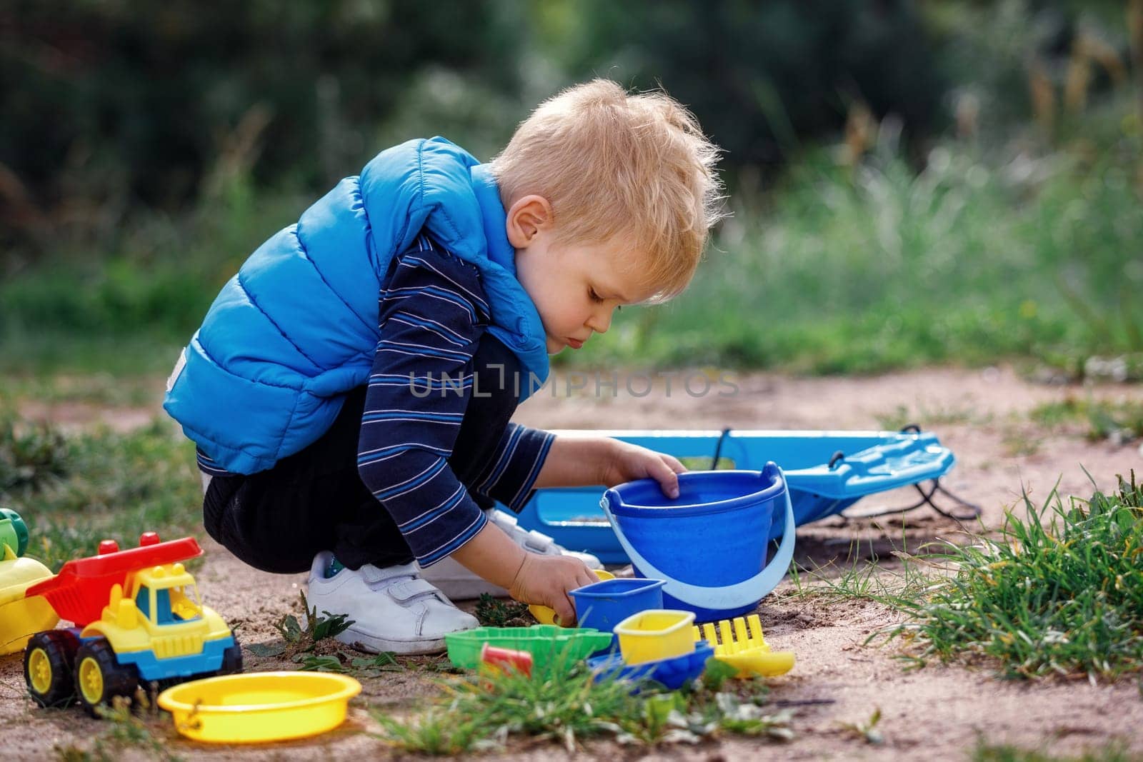 Child squat on the ground playing with sand toys. The child digs the sand into a plastic bucket. Summer outdoor activity for kids. Leisure Time.