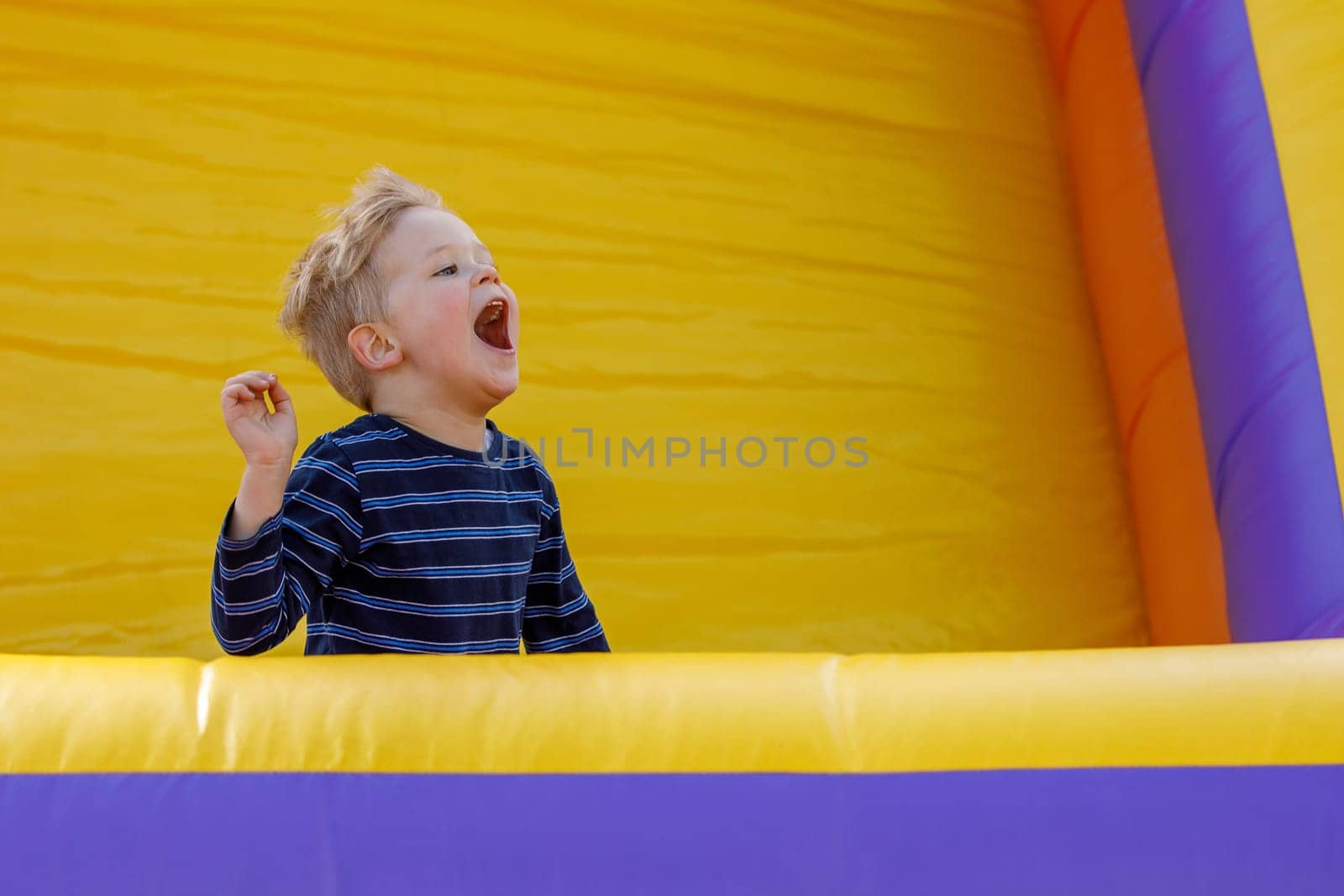 Little boy having fun in inflatable castle playground. The child is naughty and shouts loudly. Bright yellow rubber trampoline background, there is free copy space by Lincikas