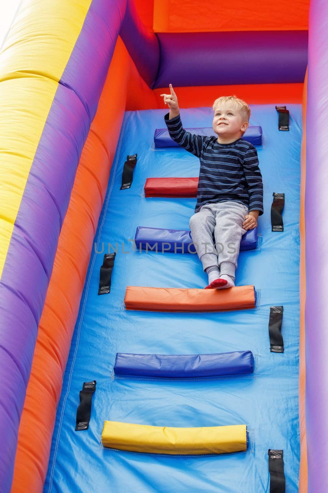 A brave and clever little sympathetic boy sits on the ladder of a large inflatable trampoline, he rejoices and watches the sky.