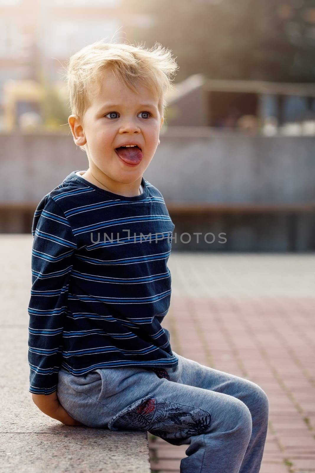 A portrait of a little naughty, sympathetic boy sitting by a city fountain, the child poses, showing his tongue.