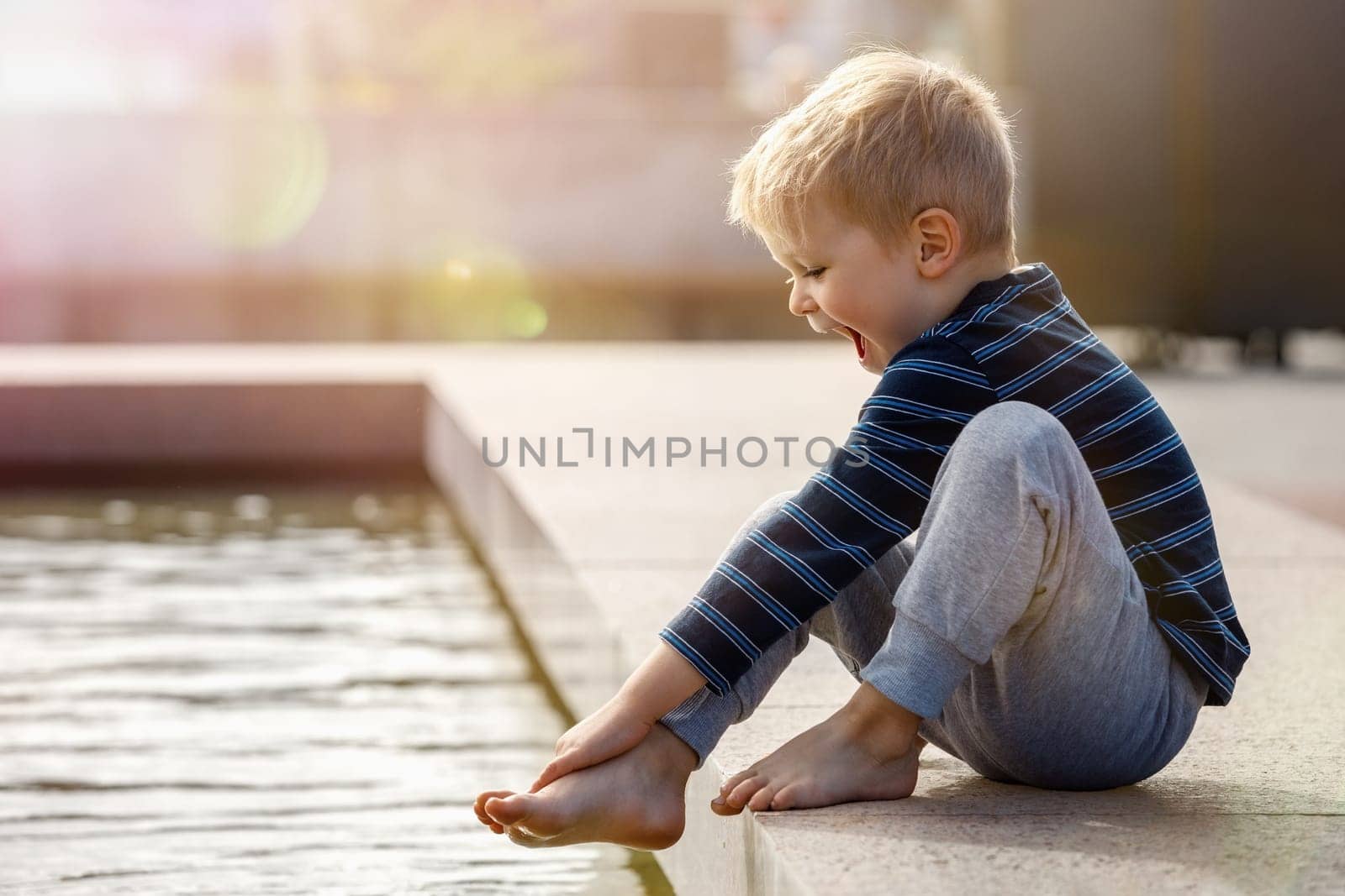 A hilarious boy on a hot summer day in the city is happy to try the cool water of the fountain on his barefoot. The child is surprised by the low water temperature.