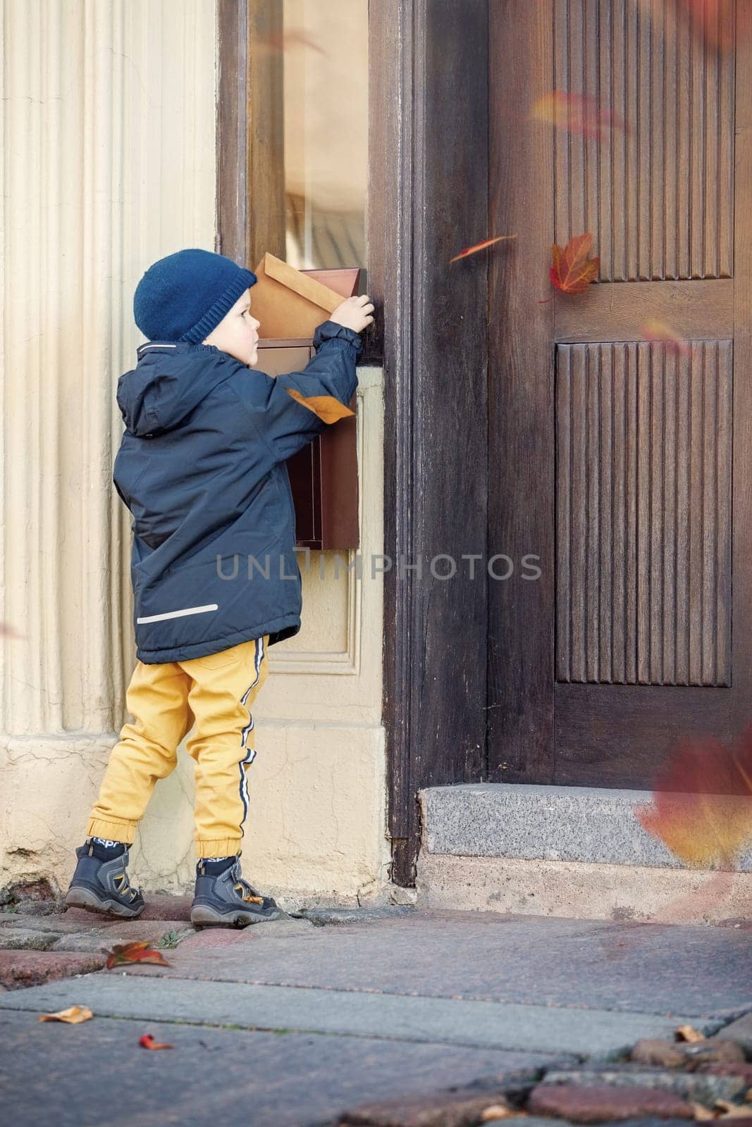 Cute boy sending or receiving mail with mailbox near house. Autumn, the wind blows colorful tree leaves.