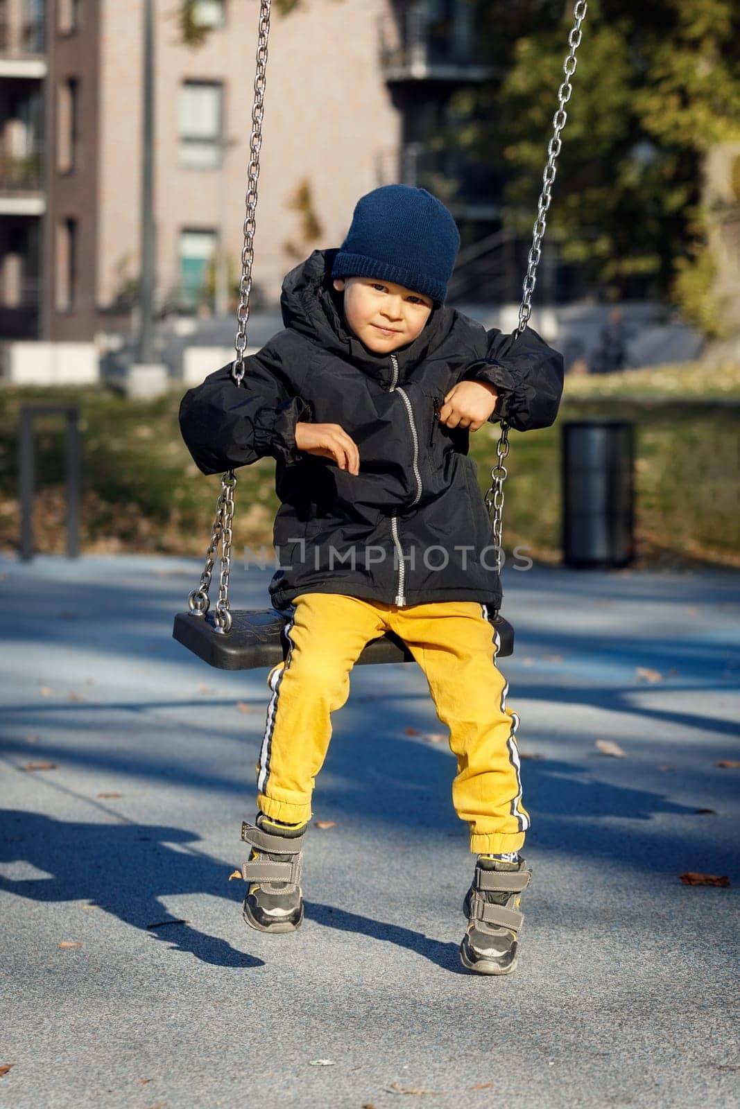 A cute boy in warm autumn clothes, yellow pants and a blue hat swings on a chain swing with a smile.