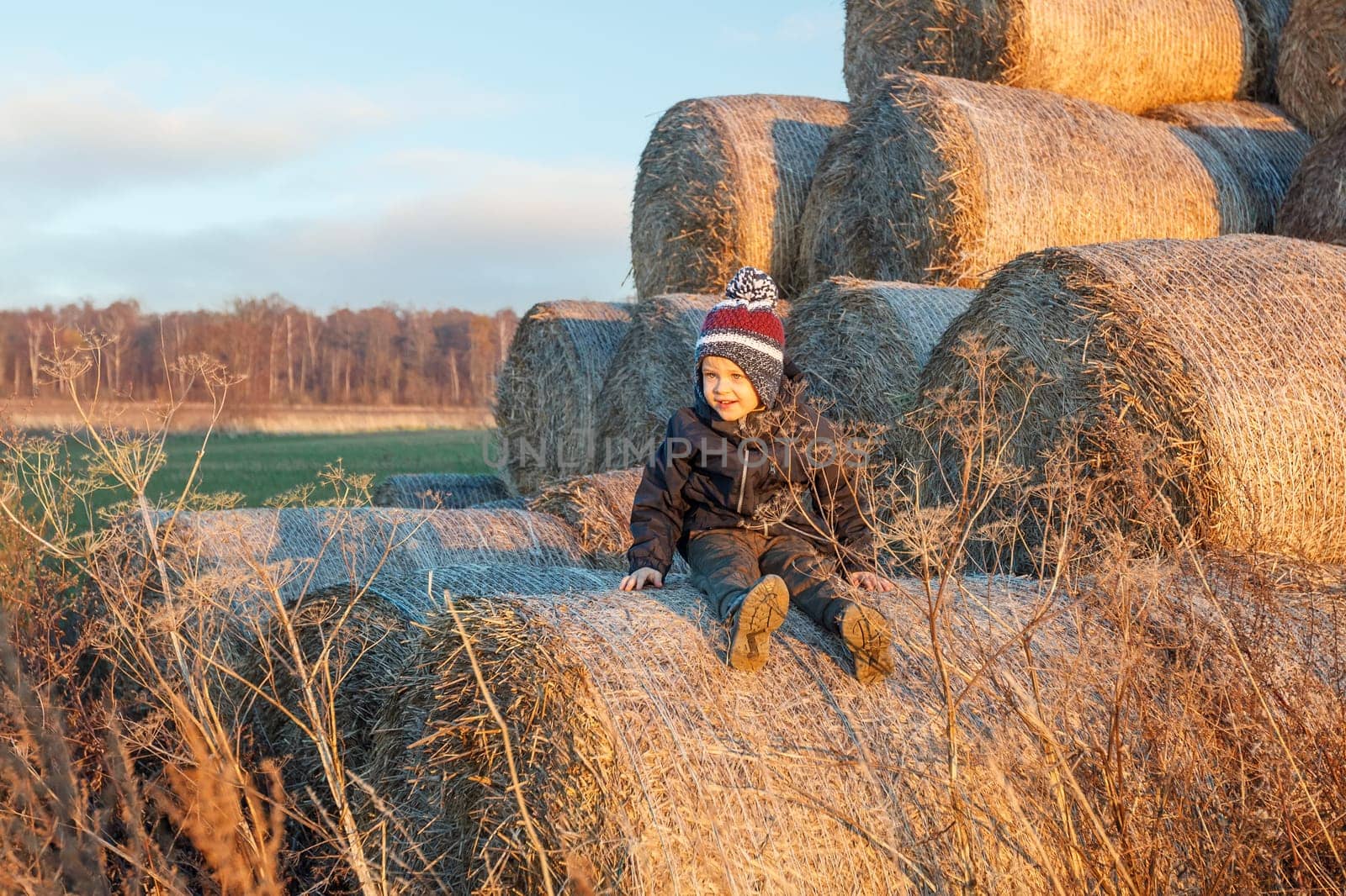 In the background of golden hay bales stack, and rural countryside landscape, poses a cute smiling boy in autumn clothes and a knitted hat.