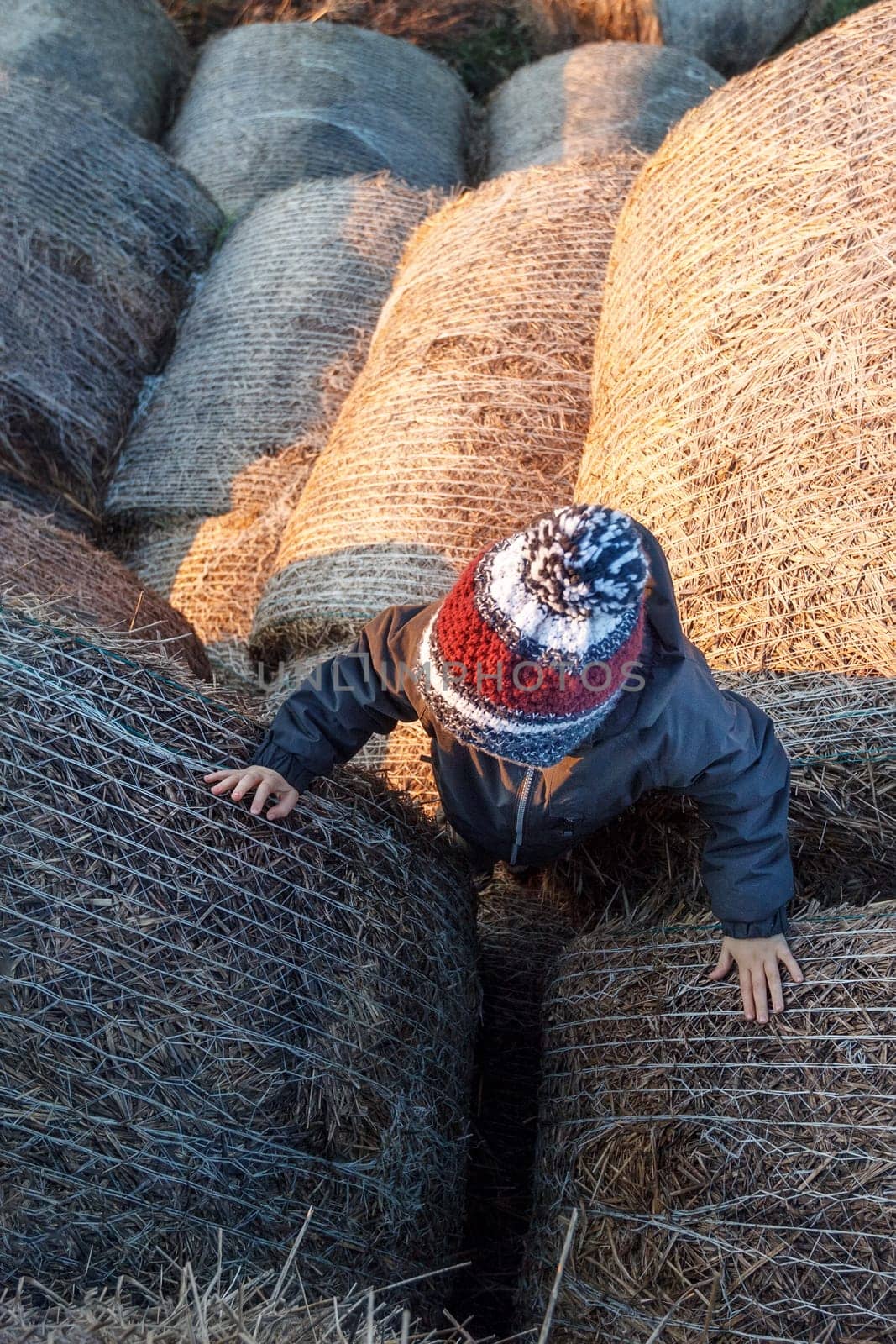 A cute boy with a knitted hat climbs to the top of a pile of straw bales.