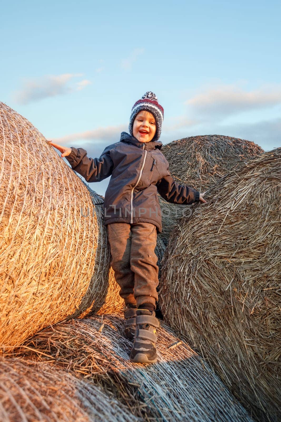 A smiling cute boy with a knitted hat stands on a golden straw stack in the evening sunlight. by Lincikas