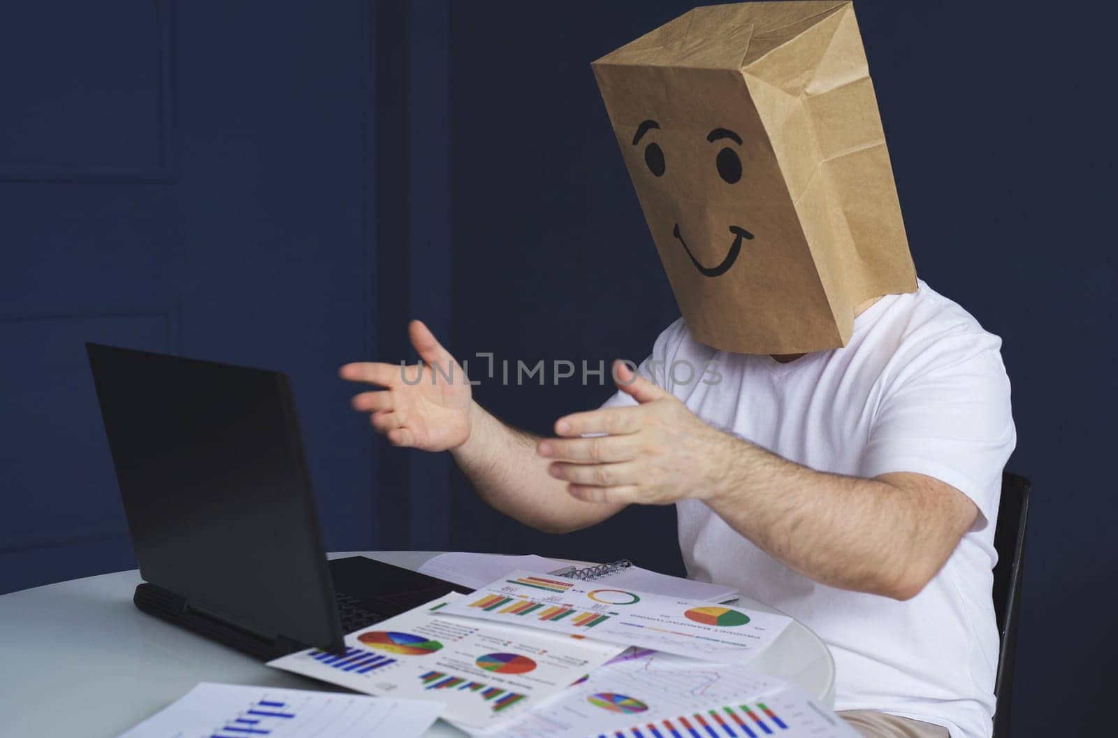 A man is a businessman in a white shirt with a paper bag on his head, with a joyful smiley face drawn, conducts a video conference or training via video link on a laptop. Emotions and gestures.