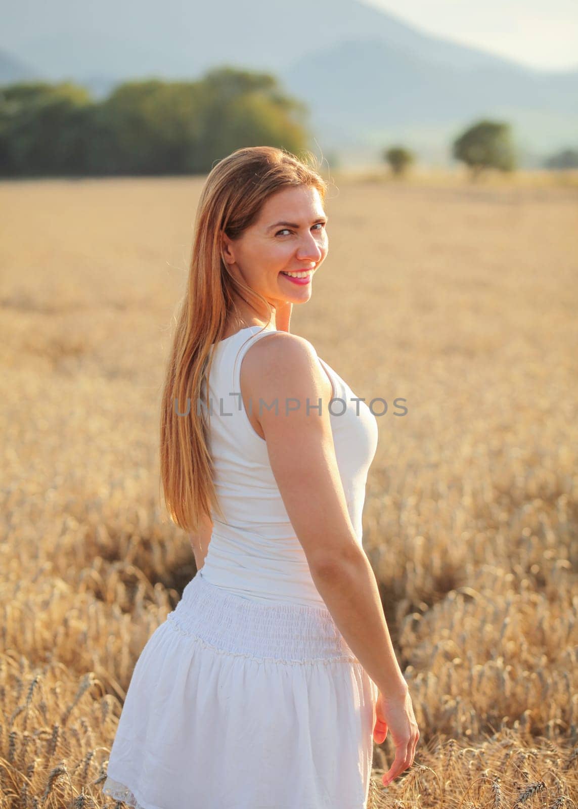 Young woman in white dress looking back over her shoulder, smiling, afternoon sun lit wheat field behind her. by Ivanko