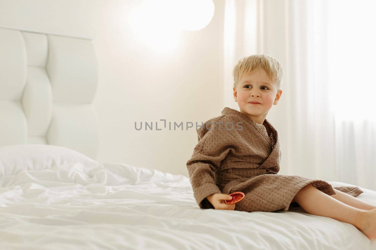 Portrait of a boy in a brown bathrobe, who is sitting on a white bed with candied oranges in his hands.