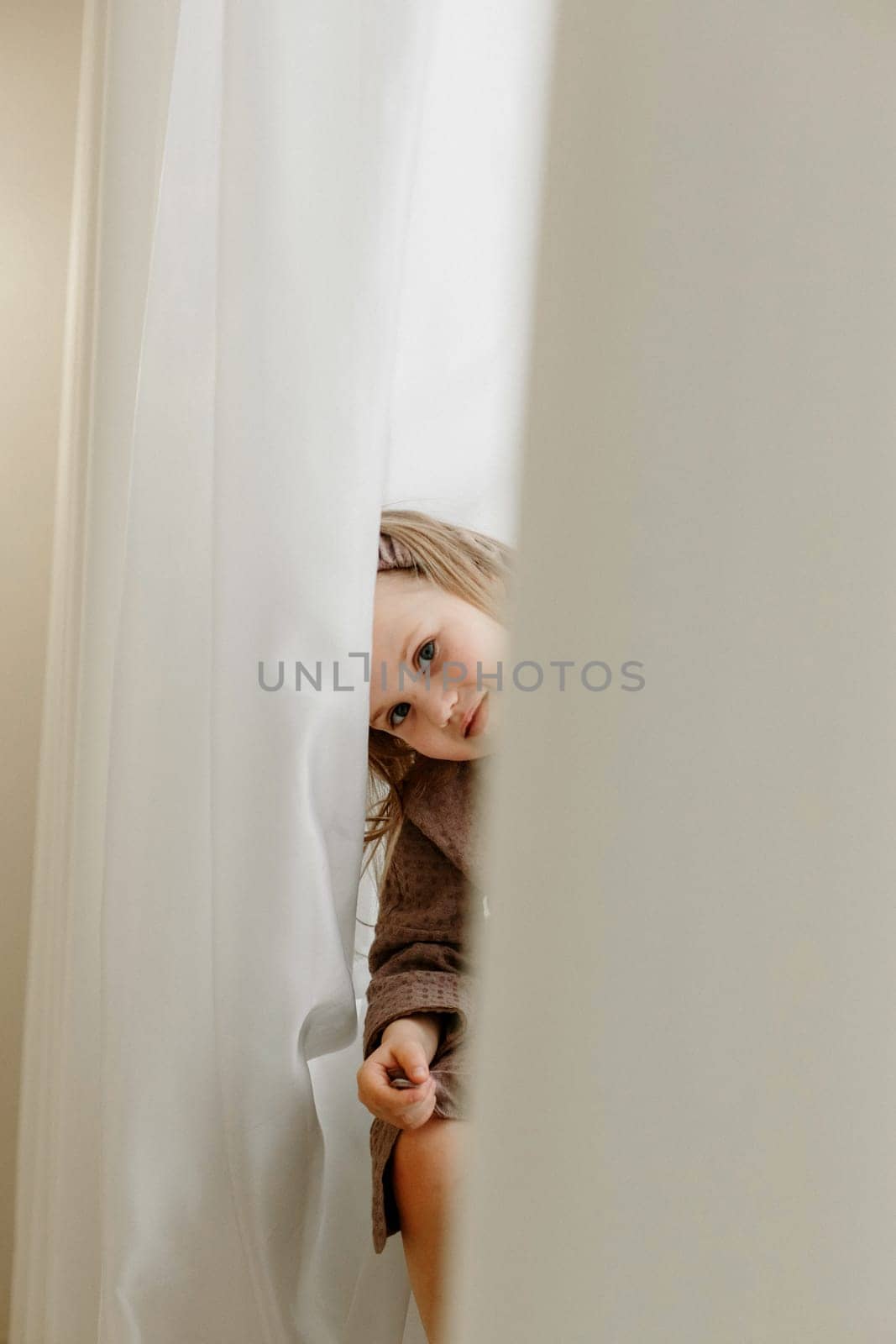 A girl in a brown bathrobe sits on the windowsill and peeps through the curtain, smiling.