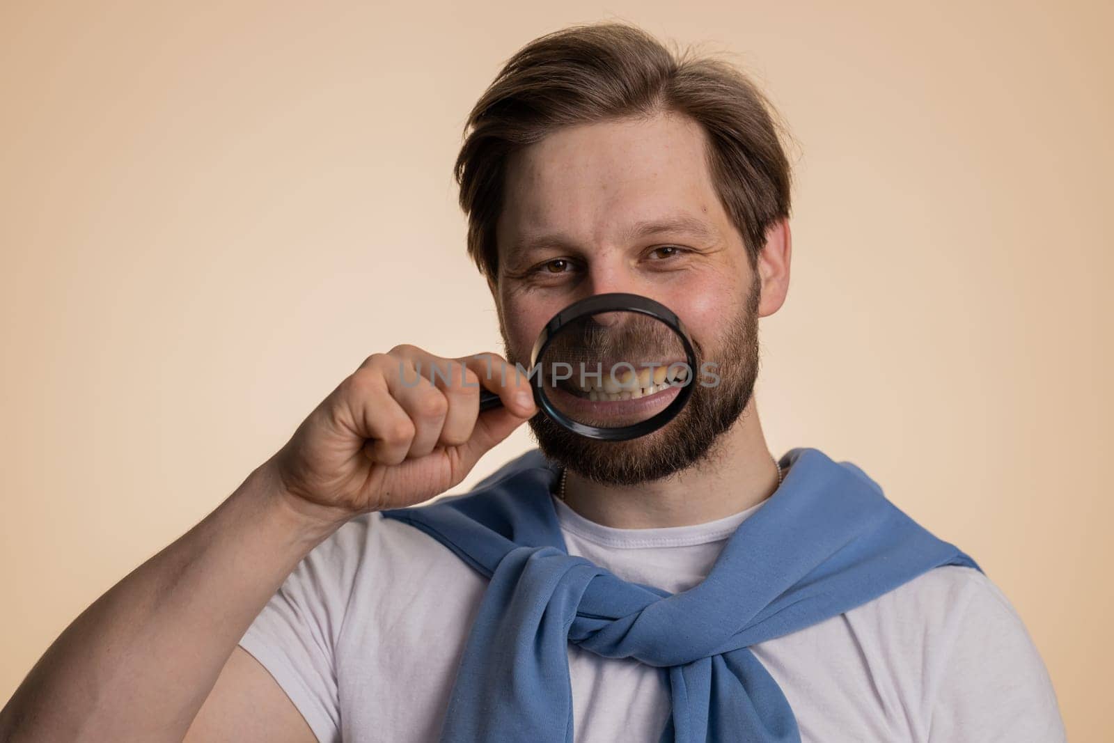 Caucasian man holding magnifier glass on healthy white teeth, looking at camera with happy expression, showing funny silly face smiling mouth. Handsome young guy isolated on beige studio background