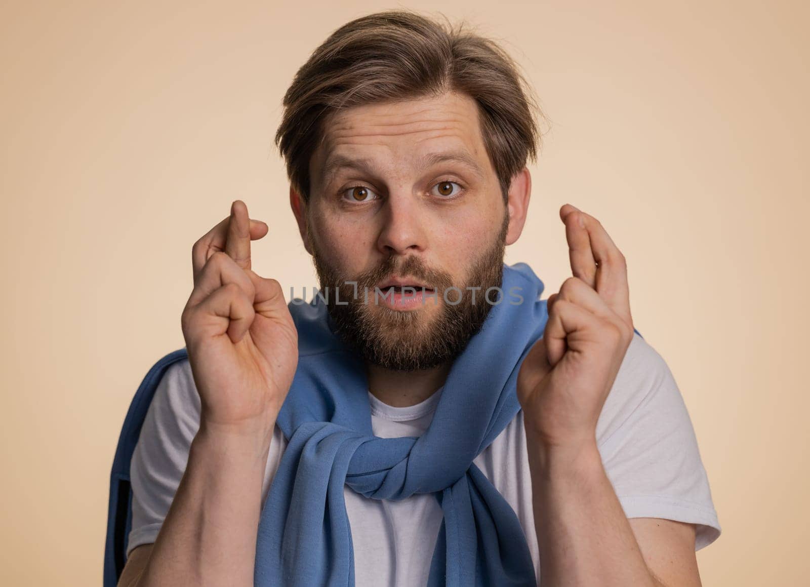 Young man crossed fingers looking at camera asking for good luck news, wishing good education exam results, dreaming about win victory in lottery jackpot. Guy isolated on beige studio background