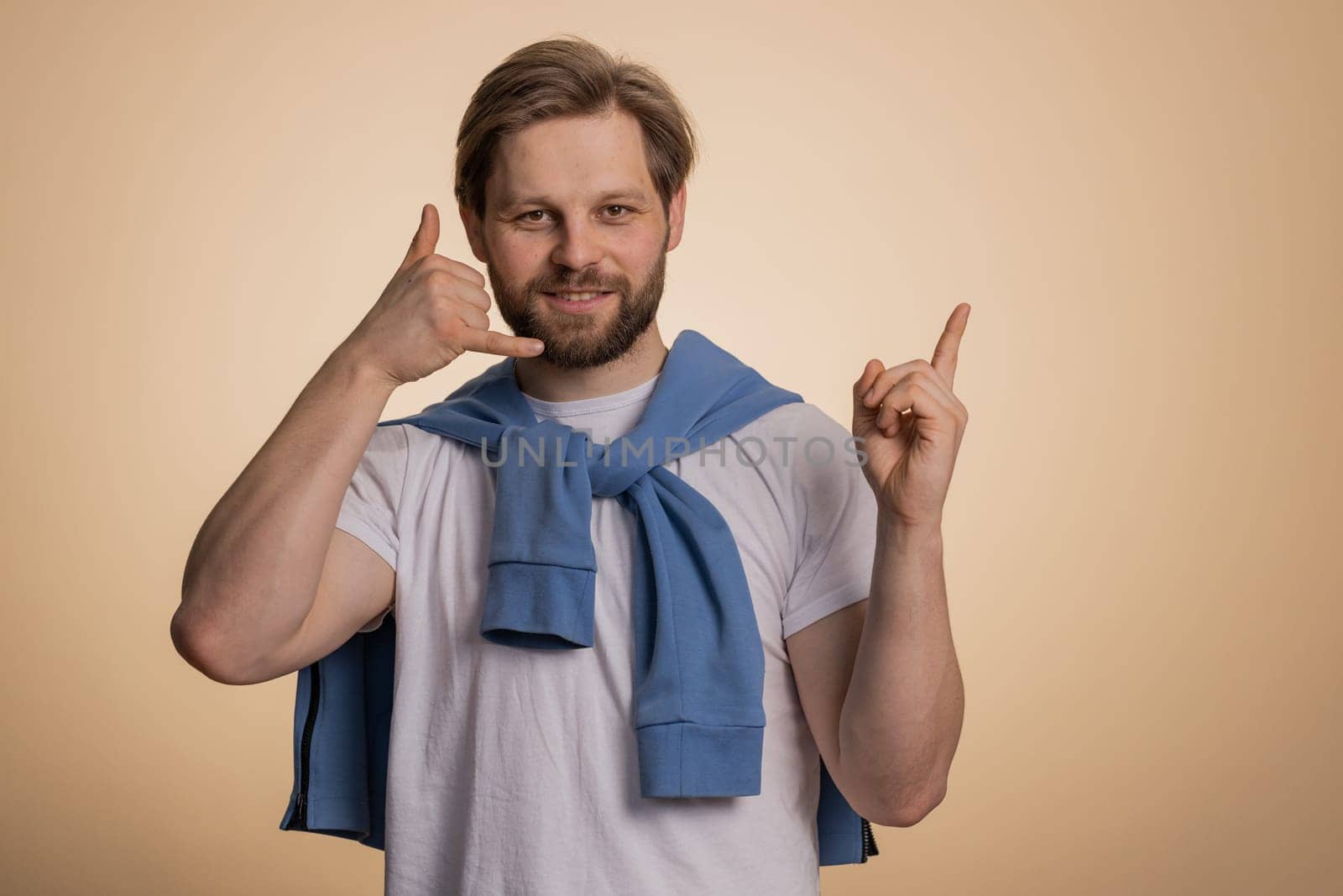 Call us, here is contact number. Handsome bearded caucasian man in t-shirt looking at camera doing phone gesture like says hey you call me back conversation. Guy isolated on beige background, indoor