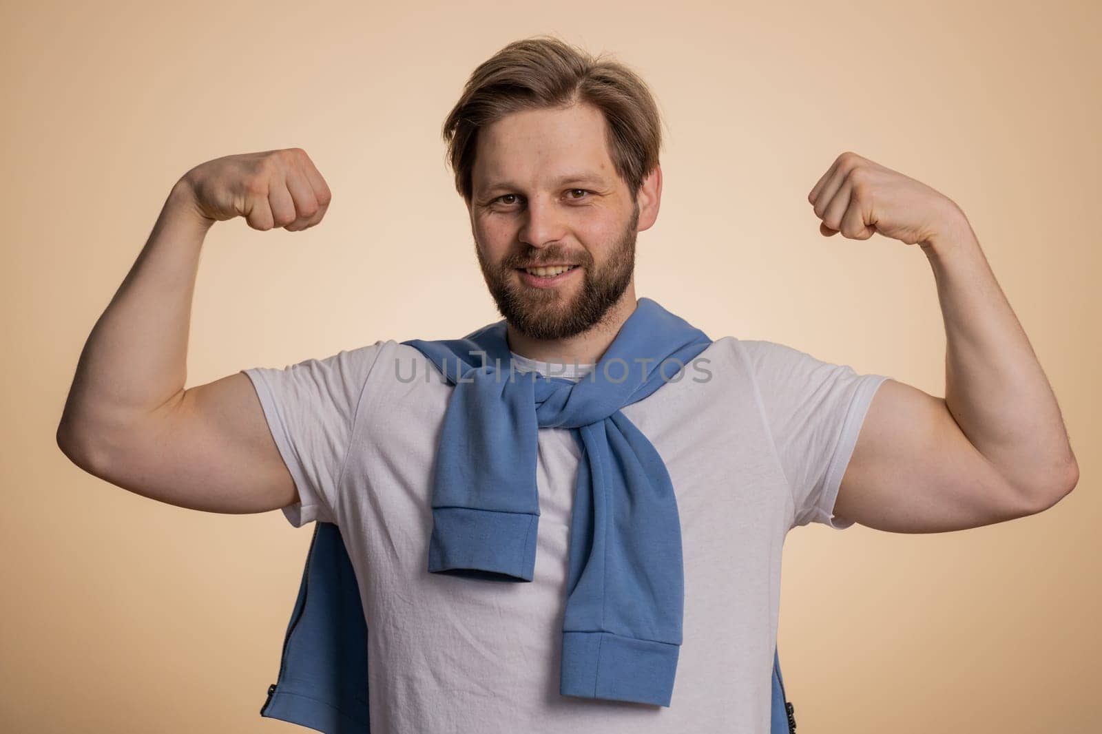 I am strong. Fit sporty caucasian man showing biceps and looking confident, feeling power strength to fight for rights, energy to gain success win. Guy isolated on studio beige background indoors