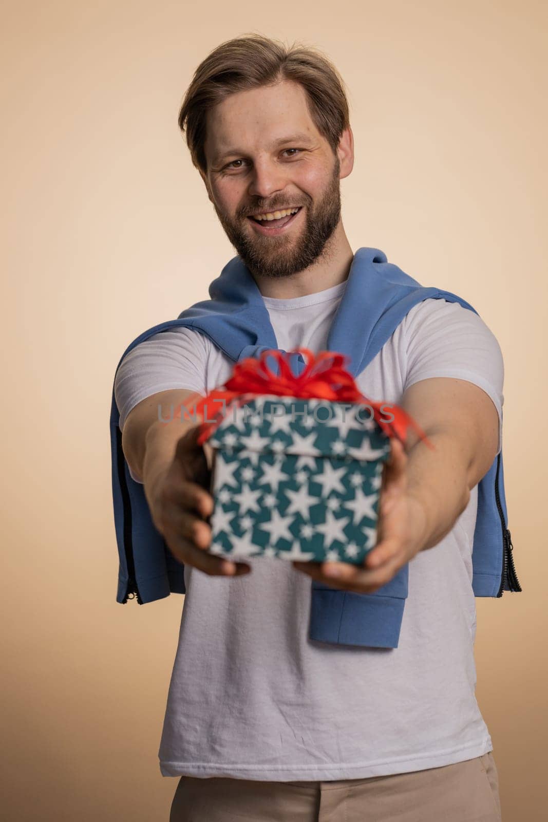 Positive smiling young man presenting birthday gift box stretches out hands, offer wrapped present career bonus, celebrating party, sale. Bearded Caucasian guy isolated on beige studio background