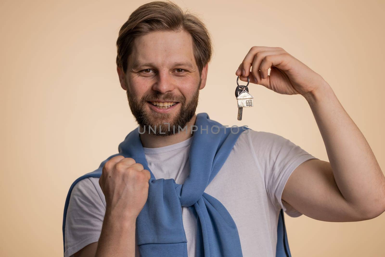Happy caucasian man winner real estate agent lifting hand showing the keys of new home house apartment, buying or renting property, mortgage loan. Young bearded guy isolated on beige studio background
