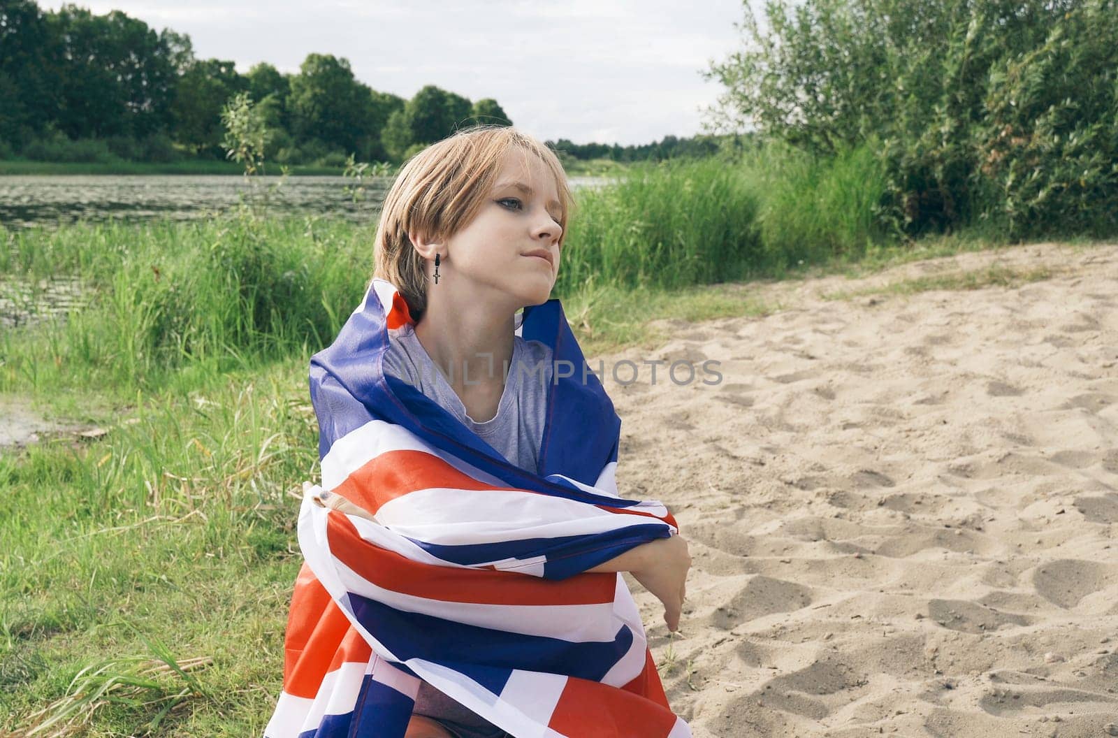 Cheerful beautiful young girl is sitting in the park hiding herself with the flag of Great Britain.