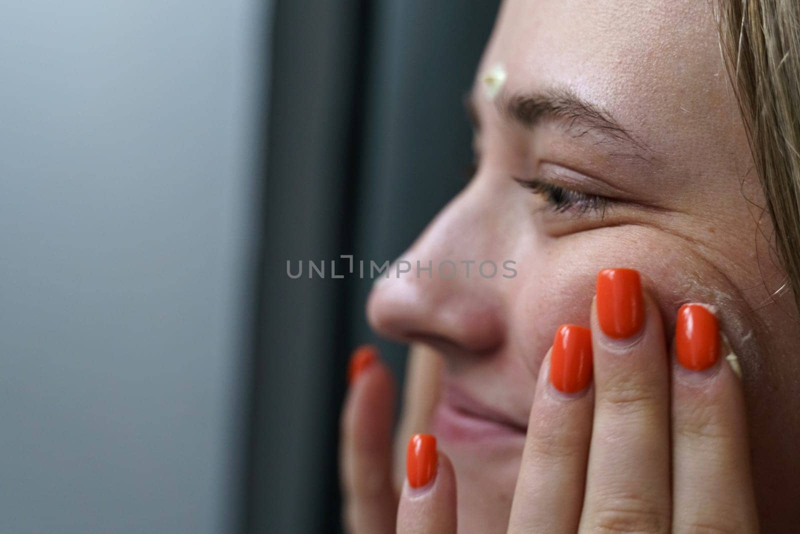 A young happy woman takes care of her face in the morning. Massages the face with hands, evenly distributing the cream over the skin. Image partially out of focus. Close-up