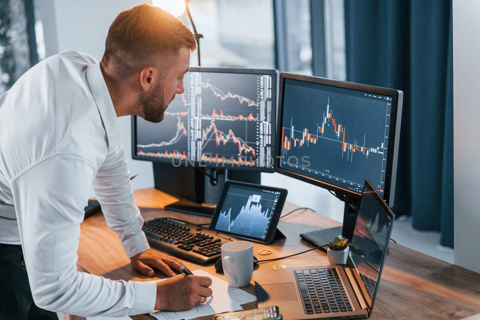 Near the table. Young businessman in formal clothes is in office with multiple screens.